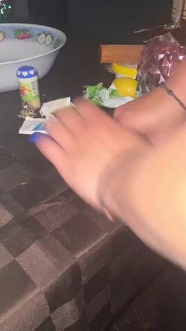 The video shows a close-up view of a person's hands rapidly slapping and tapping a dark brown and black checkered tablecloth. The activity appears to be a rhythmic hand-slapping game or percussive exercise. On the table, several objects are visible: a white bowl with a patterned rim, a blue and white cylindrical object, a pack of "OMNI" cigarettes, a blue lighter, small green wrappers, a yellow fruit-shaped object, and a pink faceted decorative item. A light blue smartphone is partially visible on the left side of the table. The person wears black bracelets on both wrists. The background is dark and somewhat out of focus, with glimpses of what appear to be decorative lights. The artificial lighting suggests it is likely evening or nighttime. The location is Belopoptsi, Bulgaria.