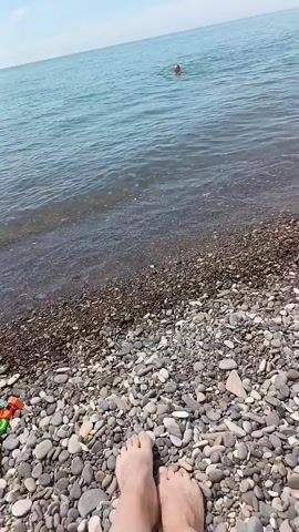 The video shows a first-person view of bare feet resting on a pebble beach, with the ocean in front. Small waves lap the rocky shore. In the mid-ground, a pair of yellow Crocs sits on the pebbles. Further out in the calm, blue-green water, a person is swimming, and an inflatable red paddleboard or kayak is visible closer to the right side of the frame near the shore. Distant structures, possibly construction equipment, are faintly visible on the horizon to the far right. The sky is clear blue with minimal clouds.