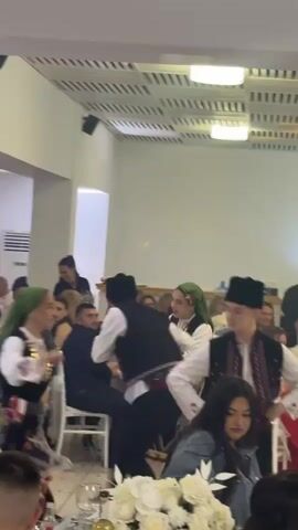 In a brightly lit reception hall, several people dressed in what appear to be traditional folk costumes, including black vests, white shirts, and colorful skirts and headdresses, perform a circle dance for an audience seated at round tables adorned with white flower centerpieces. Many audience members watch with smiles, and some take photos or videos. The room has white walls, light-colored flooring, and a decorative ceiling with recessed lighting. An air conditioning unit is visible in the background. The overall atmosphere seems celebratory and festive.