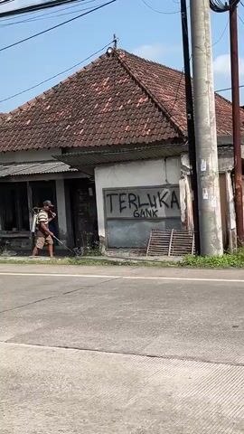A man is spraying something from a backpack sprayer in front of a small, single-story building. He is wearing a striped shirt, shorts, a hat, and a mask. The building has a tiled roof and a corrugated metal awning. There is graffiti on the wall that says "TERLUKA GANK". The scene appears to be outdoors in a rural or suburban area, possibly in Indonesia, given the location context. The time of day seems to be daytime, likely morning or afternoon, given the bright sunlight and clear sky. The man is focused on his task, and there are no other people visible in the immediate vicinity. The building looks like a shop or a house with a storefront. There are some wooden structures and stacked items outside the building. The road in front of the building is paved.