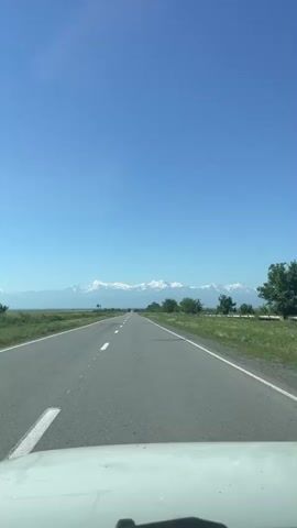 The video shows a view from the perspective of a person in a vehicle traveling on a paved road. The road is straight and appears to be in good condition, with a clear white line dividing the lanes. On either side of the road, there is green grass and some scattered trees. In the distance, a range of snow-capped mountains is visible against a clear blue sky. The sun is shining, and the overall impression is of a bright, sunny day in a scenic, open landscape.