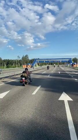 The video shows a group of motorcyclists riding on a multi-lane highway on a clear day with fluffy white clouds in a blue sky. The camera is mounted on a motorcycle in the foreground, capturing the perspective of one of the riders. In the distance, there is a pedestrian overpass with blue panels and signs displaying "90", indicating a speed limit. The highway has clear lane markings and some roadside barriers. Other vehicles, including a truck, are also visible on the highway. The motorcyclists appear to be traveling in formation, with the camera motorcycle in the lead or close to it. The environment seems to be a rural or semi-rural area, with trees visible beyond the highway.