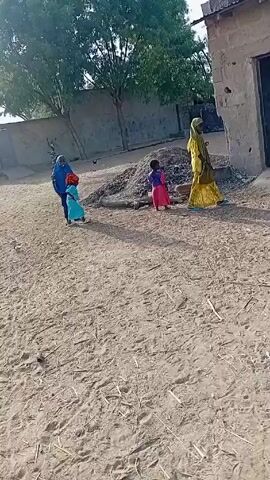 The video shows a dirt yard outside a simple building. Three children, each wearing brightly colored traditional clothing, are walking and dancing on the dirt, with an adult woman in a yellow dress watching from the doorway of the building. The yard is dry with scattered debris. Another building and some trees are visible in the background. The children appear to be playing or mimicking dance steps. The camera is handheld and moves slightly, following the action.