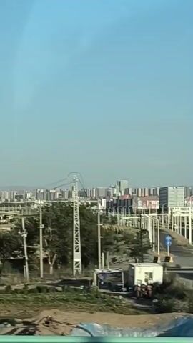 The video captures a view from a moving vehicle, likely a train or bus, looking out at a cityscape. The scene is dominated by a clear blue sky and a wide road with multiple lanes lined with streetlights. Buildings of varying heights, some appearing to be modern high-rises, are visible in the distance, forming a dense urban skyline. There is some greenery with trees and a grassy area along the side of the road. A few cars are seen on the road, and there are road signs. In the foreground, a section of the vehicle's interior railing is visible. The overall impression is of a typical urban or suburban environment.
