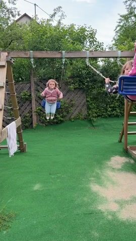 The video shows two young girls on swings in a backyard setting. The swings are part of a wooden playset that includes a ladder and rope climbing feature. The ground beneath the playset is covered with green artificial turf. One girl is jumping on her swing, while the other is seated and swinging. Two adults are present; one is filming the children with a smartphone, and the other is visible briefly behind the ladder. The yard is enclosed by a wooden fence, with trees and a building visible in the background. The sky is overcast.