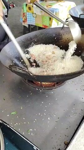 A street food vendor uses two large cooking utensils to toss fried rice in a black metal wok over an open flame on a metal work table. A small, checkered-covered table sits in the background with people seated at it. A few grains of rice are scattered on the countertop.