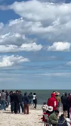 Certainly! Here's a description of the video:

**Scene:** The video captures a group of people gathered on a sandy beach, watching a formation of military aircraft fly across the sky. 

**Environment:** The setting is a sunny day with a bright blue sky scattered with puffy white clouds. A body of water (presumably the ocean or a large lake) stretches to the horizon. The beach itself is sandy, and the light suggests it's a clear day.

**People:** There is a crowd of people of mixed ages and clothing, standing or sitting on the beach, facing the water. Most of them appear to be focused on the sky, likely observing the aircraft.

**Objects:** The primary object of interest is the formation of aircraft. There appear to be 5-6 of them flying in a precise arrangement against the bright blue sky. A few beach chairs are visible on the sand.

**Actions:** The main action is the flight of the aircraft, which is capturing the attention of the people on the beach. The people themselves are standing and looking up, some may be taking pictures or videos with their phones.

**Overall Impression:** The video conveys a scene of excitement and anticipation, with people gathered to witness an impressive display of aerial maneuvers.