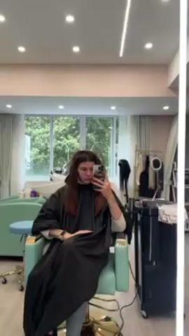 A woman is in a hair salon and taking a selfie. She has brown hair with foils placed in sections at the top of her head. She is wearing a black salon cape and sitting in a turquoise chair. The salon has a large window with a view of trees. There is hair styling equipment on a rolling cart to the right of the chair. The room has a light, neutral color scheme.