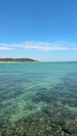A vertical shot shows a clear turquoise sea with a rocky seabed visible beneath the surface. In the distance, a hazy coastline with green hills and a crowded beach can be seen under a bright blue sky with scattered white clouds. Several small boats and people swimming in the water are also visible. The water is calm with gentle ripples on the surface.