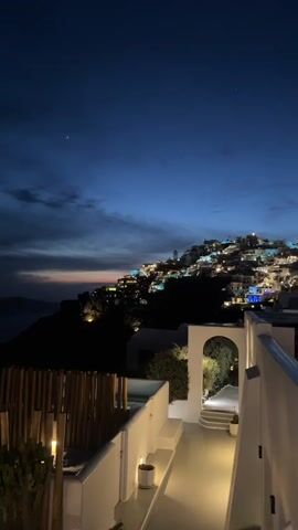 The video shows a night view of a hillside village with white buildings, likely in Greece. The sky is a deep blue with some wispy clouds and a visible star. The village is illuminated with warm lights. In the foreground, there's a white terrace or balcony with a bamboo screen and an arched opening leading to a pathway. The scene is still and quiet, emphasizing the tranquility of the location.
