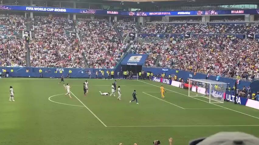A soccer match is underway in a packed stadium. A player in white is down on the field, appearing to be injured, while other players in white and blue surround him. The goalkeeper is near the goal. The stadium is filled with spectators, and banners overhead indicate the event is the FIFA World Cup 2026 Bronze Final in Miami.