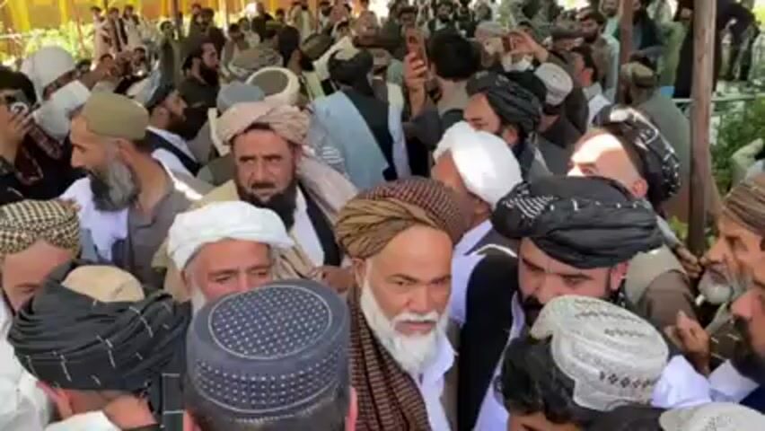 Here's a concise, forensic description of the video:

The video shows a densely packed crowd of men, predominantly wearing traditional Afghan clothing, including turbans of various colors and patterns. Many have beards, and their faces show a range of expressions, some appearing somber. The environment appears to be outdoors, possibly under some sort of awning or partial shelter, suggested by varying light conditions. The background is blurry with hints of trees and other people. There is a general feeling of crowding and potential tension within the group, although specific actions or direct interactions are difficult to discern due to the close proximity of the individuals. Some individuals are using mobile phones, possibly to take photos or videos.
