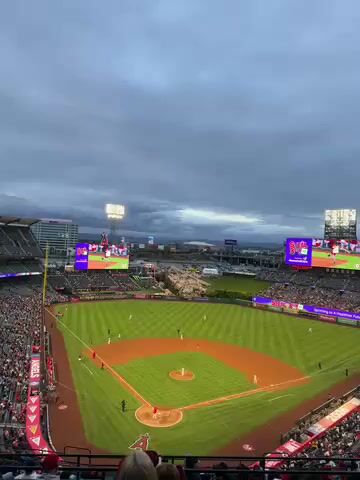 The video captures a wide, elevated view of a baseball stadium during a game. Thousands of spectators fill the stands, predominantly wearing red and white colors. On the green field, baseball players are spread across the diamond and outfield. Large stadium lights illuminate the scene under a heavily overcast and cloudy sky, suggesting either dusk or a dark daytime. Two massive video screens in the outfield display game information and the team's "A" logo. A distinctive rock formation, characteristic of Angel Stadium in Anaheim, is visible beyond the center-field fence. The sounds of an announcer and crowd engagement can be heard, indicating an active sporting event. The "A" logo is clearly visible on the scoreboards and stadium structures.