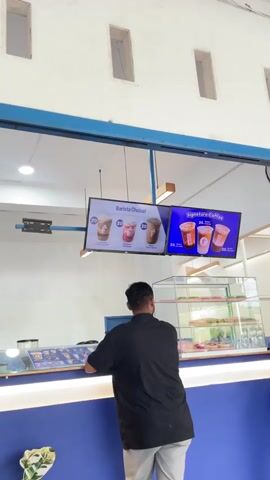 A man in a black shirt and gray pants stands with his back to the camera, looking at a menu board in a cafe. The menu board displays images of coffee drinks. To the right, a woman in a striped shirt and black pants with a hijab also looks at the menu. The cafe has a blue counter with glowing lights. Behind the counter, glass display cases show donuts. The walls are white with rectangular openings near the ceiling.
