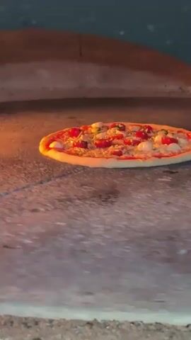 A pizza is baking in a wood-fired oven. The pizza has red sauce, white cheese, and some red toppings. The oven floor is made of stone and is warm from the fire. Flames are visible in the background. The camera is focused on the pizza as it bakes.