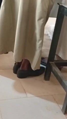 A camera initially points down at a person's feet, shod in black shoes and wearing a light tan robe-like garment. The camera then angles up, revealing a man in the same garment, who turns to face the camera and smiles. The background includes other people, a white board, and a metal table.