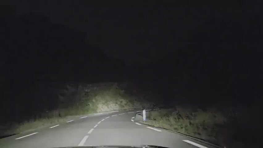 This video shows a first-person perspective of driving a car on a dark, winding road. The headlights illuminate the asphalt road with white lines. On the right side of the road, there is a white concrete barrier and dense vegetation. On the left side of the road, there is a steep incline covered in grass and rocks, leading up to a dark hillside. The sky is completely black, indicating it is nighttime.