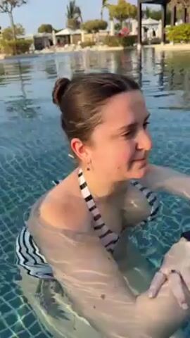 A woman in a black and white striped bathing suit stands in a swimming pool on a sunny day. The pool has a blue tiled bottom, and the water is clear. The woman is smiling at the camera. She slowly lowers herself to float on her back in the water. In the background are trees, buildings, and lounge chairs.