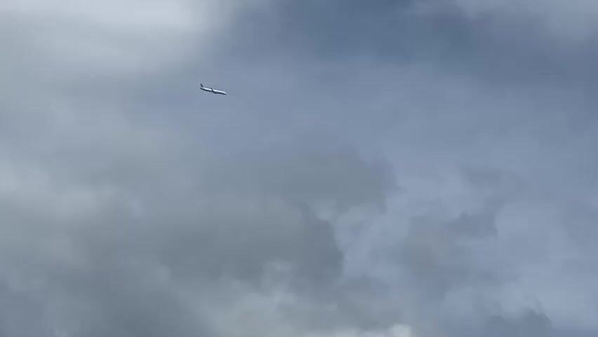 A commercial airplane is visible in the sky, flying from left to right. The sky is overcast with clouds, some of which are dark and appear to be rain clouds. The airplane is white with a red stripe on its tail. It is at a high altitude and appears to be in a stable flight.