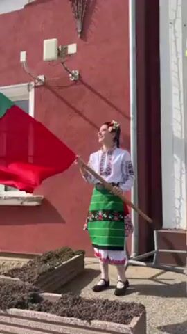 A young woman, dressed in traditional Bulgarian clothing, is holding a Bulgarian flag on a pole. She is standing in front of a red building with a window and white trim. A small green trash can is beside her and she is standing on a raised planter box. She smiles as she waves the flag.