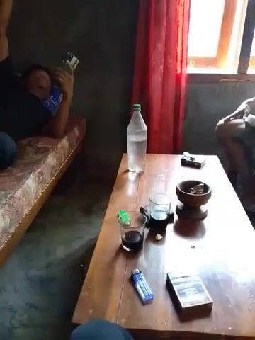 The video shows a dimly lit room with two people. One person is lying on a mattress on the floor, holding a phone to their ear. A wooden table is in the center of the room, with a bottle of water, two glasses, a cigarette pack, a lighter, and an ashtray on it. Another person is partially visible sitting on the right side of the frame. Red curtains hang in the background by a window. The lighting suggests it may be daytime, but the interior is dark.