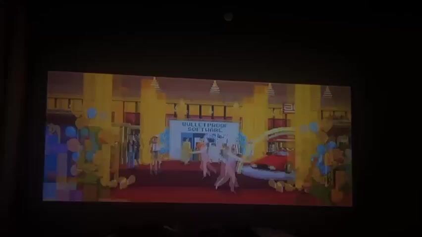 The video shows a projected image of a pixelated video game on a wall. The initial scene depicts a grand opening of a software company named "Bulletproof Software." The scene features pixelated characters, including businesspeople and bunny girls, outside the company entrance. The scene transitions to a menu screen where a character named "Henk Rogers" is identified as Player 1.
