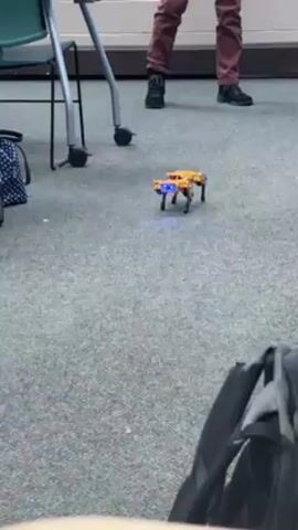 The video shows a small, robotic dog-like device walking across a carpeted floor. The robot is yellow and black with blue lights illuminating its underside. Behind the robot, an individual wearing maroon pants and black shoes is standing. In the background are chairs and a backpack, providing a classroom or similar setting context. The robot's movement is the primary action, and the presence of a person suggests observation or control of the robot.