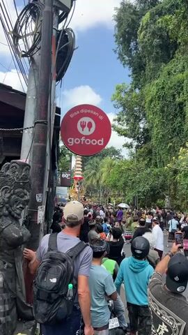 The video shows a busy street scene, likely in a tropical location, with a crowd of people gathered. Overhead, a red circular sign with a white fork and knife icon and the text "gofood" is visible. Below the sign, a tall, ornate structure, possibly a ceremonial tower, is being carried or paraded through the crowd. Several people are holding up their phones, presumably to take pictures or videos. Trees and lush greenery line the street on the right side of the frame. A stone statue is partially visible on the left. The sky is overcast with some blue patches. The overall impression is of a lively event or festival.