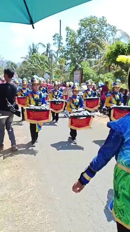 A marching band parades down a paved road under a bright sky. The band members, all young people, wear matching blue and gold uniforms with elaborate hats. They are playing xylophones, which are mounted on red banners with gold trim. The banners read "LASKAR SYMPHONY SADULUR" in gold lettering. People are watching the parade from the sides of the road. Trees and buildings are visible in the background. A large black umbrella is partially visible at the top left of the frame.