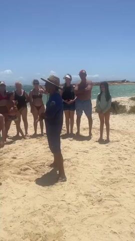 The video shows a group of about a dozen people on a sandy beach. The people are dressed in a variety of beach attire, including swimsuits, shorts, and t-shirts. An adult male, wearing a straw hat and a dark blue uniform-style shirt, stands in the center of the group and appears to be speaking to them. Most of the other people are facing the man as if listening to him. Behind the group, there is the ocean with white-tipped waves and a clear blue sky. In the background, there appears to be a shoreline with some vegetation. Other people are walking on the beach, and a boat is seen on the horizon. The lighting suggests it is a bright, sunny day.