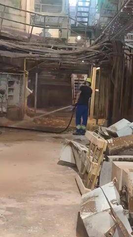 A person wearing a hard hat and blue overalls is power washing a floor in what appears to be an industrial setting. The space is dimly lit and dusty, with remnants of equipment and debris scattered around. Wooden pallets are stacked to the right, and large, metallic structures are visible in the background. The person is holding a hose, directing a stream of water onto the ground, suggesting a cleaning or maintenance activity. The overall impression is one of ongoing work in a somewhat derelict or under-construction industrial environment. No text is visible in the media.