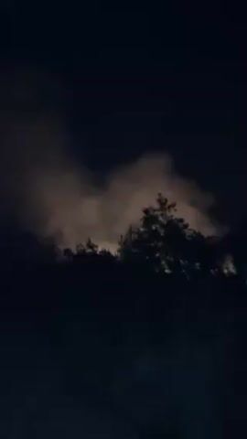 Here is a description of the video:

The video begins with a dark, low-angle shot of tree silhouettes against a sky illuminated by what appears to be a yellowish light, possibly from a fire or bright light source obscured by clouds. The camera then shifts downward, providing a close-up view of the ground and the legs of a person wearing blue jeans and dark shoes. The person seems to be walking on a paved surface. The camera angle is low, suggesting it is attached to the person or held close to the ground. The overall impression is of someone moving through a dark environment with an ominous glow in the background.