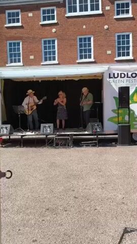 A band performs on a trailer stage in front of a brick building with white-trimmed windows. The stage banner reads "LUDLOW GREEN FESTIVAL." Three musicians are visible. A man on the left plays an acoustic guitar and wears a light-colored shirt and a straw hat. A woman in the center plays a flute and wears a black and white polka-dot dress. A man on the right plays an electric guitar and wears a green shirt. Two speakers are positioned on the stage in front of the musicians. The ground in front of the stage appears to be gravel.