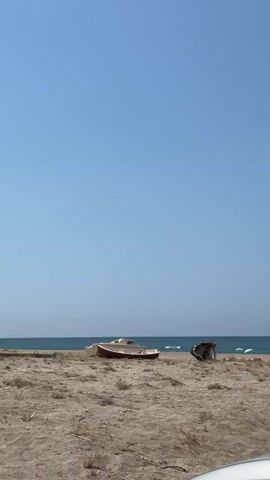 A wide shot of a beach on a clear, sunny day. The sky is a vibrant blue with no clouds. The ocean is a calm, deep blue with gentle waves breaking on the shore. In the foreground, a sandy beach stretches across the frame, with sparse, dry vegetation growing in patches. Two objects are visible on the sand: a small, overturned red and white boat, and to its right, a dark, vaguely cylindrical object that appears to be debris. The camera is stationary, capturing the static scene.