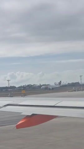 The video shows a view of an airport tarmac from an airplane window. Part of the wing of the plane, painted with an orange accent, is visible. In the distance, another airplane, painted with green, red, and white markings, is parked on the tarmac. There is a yellow sign with black numbers on it, near the parked airplane. The sky is overcast with a mix of clouds. The wing of the plane gradually reveals that it is an EasyJet airplane.