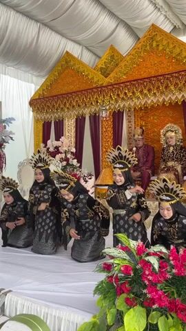 A group of five young women in black and gold traditional attire with ornate headpieces perform a dance on a white stage. They are kneeling, then rise and move their arms in coordinated gestures. In the background, a couple sits on a raised, ornate throne draped in gold and burgundy fabric. The environment appears to be an indoor event space with white draped walls and ceiling.