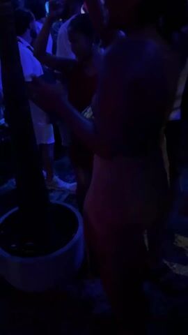 The video shows a person with their back to the camera dancing in a crowd. The individual is wearing a tight, light-colored dress with thin straps and a cutout in the back. The scene is dimly lit, with purple and blue lighting creating an ambiance. Other people in the crowd are dancing and moving around, mostly dressed in white shirts and lighter-colored clothing. There's a large potted plant on the left side of the frame. The music is loud and rhythmic, suggesting the setting is a party or club.