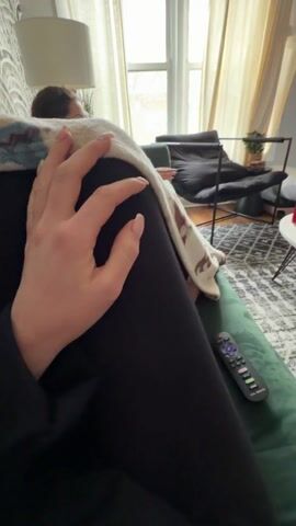 The video shows a person relaxing on a green couch in a living room. A woman is visible on the couch behind them. The living room has a grey patterned rug, a white coffee table, and a TV playing a Hannah Montana concert video. There's a black chair and a large window with beige curtains in the background. The camera pans to show the TV, which is playing a live performance with the words "Hannah Montana" displayed prominently. The person is holding a TV remote and is covered with a blanket.