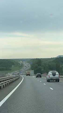 The video shows a car driving on a multi-lane highway. The road winds through green, hilly terrain under an overcast sky. Other vehicles, including cars and a truck, are visible on the highway. There is a white line marking the edge of the lane. Raindrops are visible on the windshield of the camera car.