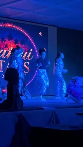 A group of children are performing a dance routine on a stage. The stage is lit with various colored lights, including purple, blue, green, and red. Behind the dancers is a large screen displaying a graphic with palm trees and stars. The dancers are wearing shimmering costumes. Some of the children are wearing silver outfits, while others are wearing colorful dresses. The children are all actively dancing and interacting with each other. The audience is not visible in the frame, but the lighting suggests a performance setting.