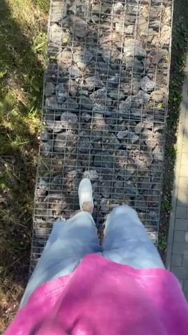 A first-person perspective video shows a person walking on a path. The path appears to be constructed from a wire mesh cage filled with rocks. The person is wearing white sneakers, light blue denim shorts, and a bright pink shirt. They are walking on the rocky path which is surrounded by green grass and foliage on the left, and a paved surface on the right. The sun is shining, casting shadows on the path. The person's legs and feet are visible as they walk away from the camera.