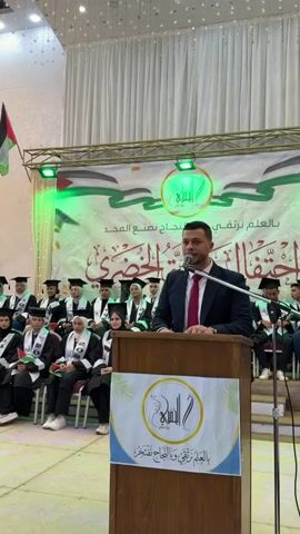 A man in a suit and tie stands at a wooden podium speaking into a microphone. Behind him, a group of people wearing graduation caps and gowns are seated on risers. A banner with Arabic script and a logo hangs behind the seated graduates. A Palestinian flag is visible in the upper left corner of the frame. The environment appears to be an indoor event space, possibly a hall or auditorium, with stage lighting and decorative elements. The man at the podium gestures with his hand as he speaks. The seated individuals are looking towards the podium, suggesting they are attending a ceremony or presentation.