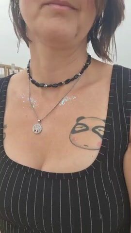 A woman stands outside on a beach. She is wearing a black pinstripe tank top, earrings, and two necklaces. A tattoo of a panda is visible on her chest.  Glitter is scattered across her chest, shoulders, and face. The beach and a structure are visible in the background.