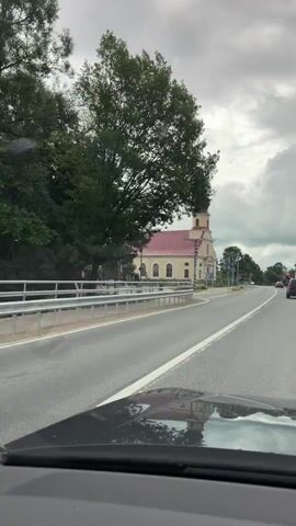 This video shows a view from inside a moving vehicle, looking forward and slightly to the right. The vehicle appears to be traveling on a paved road. To the right of the road, a light-colored church with a red roof and a dark steeple is prominently visible. The church has several arched windows along its facade. In front of the church, there is a sidewalk and a grassy area. The road has lane markings, and there are traffic signs and streetlights present. Other vehicles can be seen in the distance on the road. The sky is cloudy. The reflection of the church and the sky is visible on the vehicle's hood in the foreground.
