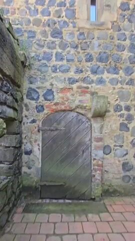 This video showcases a stone tower, likely a historical structure like a castle or fortification, in Stolpen, Germany. The tower is constructed from rough, dark stones interspersed with reddish-brown bricks, giving it a weathered and ancient appearance. Several windows punctuate the stone facade; some are simple openings with small panes of glass, while others are reinforced with metal bars, suggesting a defensive purpose. One window, in particular, is a small, rectangular opening with a stone frame.

At the base of the tower, a weathered, wooden door with diagonal planks and metal hinges stands slightly ajar, hinting at entry into the structure. The scene is shot from a low angle, looking upwards, emphasizing the height and imposing nature of the tower. The sky above is visible, with patches of blue and white clouds, suggesting daytime with fair weather. The lighting indicates it could be morning or afternoon.

The overall impression is one of history and resilience. The rough texture of the stones, the aged wood of the door, and the secure windows all speak to the tower's past as a functional building, possibly a defensive stronghold. The moss and greenery growing on parts of the stone and around the base add to the sense of age and the way nature is reclaiming the man-made structure. There is no visible text in the video.