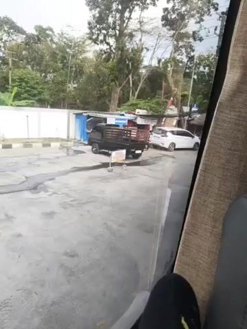 The video shows a view from inside a vehicle, looking out through the windshield. The scene outside appears to be a roadside area with a few small structures and a paved lot. In the foreground of the lot, there is a black pickup truck with wooden sides, parked facing towards the left. A yellow sign is placed in front of the truck. To the right of the truck, a white car is parked. Further in the background, there are trees and some overhead wires. Another white van is visible in the distance, moving away. The overall environment suggests a rural or semi-rural setting, possibly a small market or roadside stop.