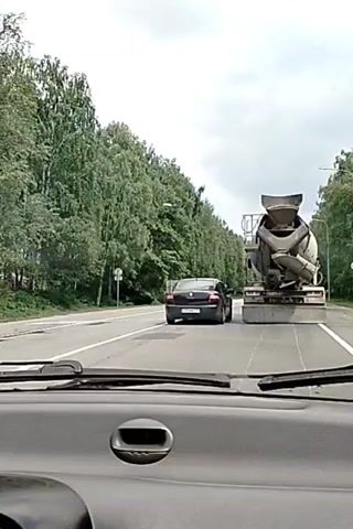 A dashcam view shows a person driving on a road lined with trees. Ahead of the vehicle, a black car and a concrete mixer truck are driving in the same lane. The concrete mixer truck is larger and appears to be moving slower than the black car. The road is paved and appears to be in good condition. The sky is overcast.