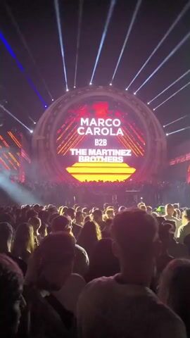 A concert is in progress. A large, circular screen displays the names "MARCO CAROLA B2B THE MARTINEZ BROTHERS." Spotlights shine from above and beams of light project from the screen. A large crowd of people is gathered in front of the screen. The crowd is mostly obscured by darkness, but some individuals are visible in the foreground and mid-ground. The people appear to be watching the performance, with some raising their hands. The overall atmosphere is energetic and celebratory.