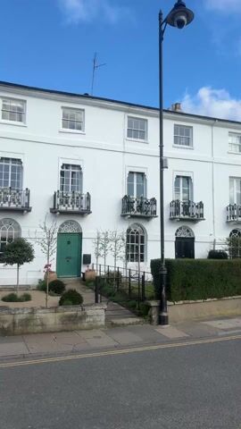 The video shows a street view of a row of terraced houses on a sunny day. The camera pans from right to left, showcasing the architectural style of the houses. The first building is painted white with a green front door and wrought iron balconies. The rest of the houses in the terrace are painted in a similar style, and have gardens with hedges and trees in front of them. The street in front of the houses has double yellow lines and several cars parked along it. The sky is blue with some white clouds.