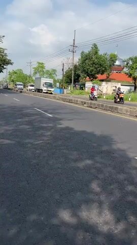 The video shows a road with a slight curve to the left, indicating a gentle turn. On the right side of the road, there is a raised concrete barrier separating the road from the embankment. Beyond the barrier, there is a sparsely vegetated area with some trees and a building with a dome, possibly a mosque. The sky is partly cloudy. Several vehicles are visible on the road, including a white truck in the foreground and two smaller white vans further down the road. The road surface is asphalt and appears to be in good condition. The lighting suggests it is daytime.